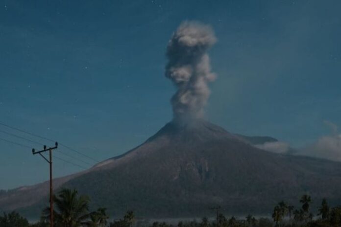 Erupsi Gunung Lewotobi memuntahkan kolom abu vertikal ke langit malam; siluet lereng gunung, pepohonan, dan tiang listrik tampak di latar depan.