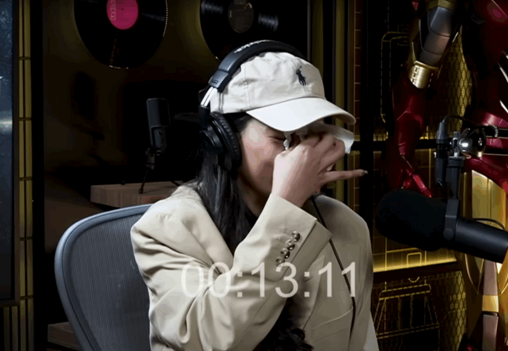 Erika Carlina menangis sambil menyeka air mata di podcast, mengenakan blazer krem, topi Polo, dan headset studio.
