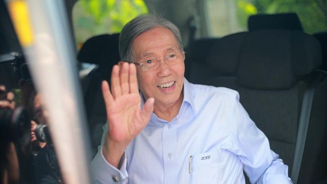 Kwik Kian Gie melambaikan tangan dari dalam mobil, mengenakan kemeja biru muda.