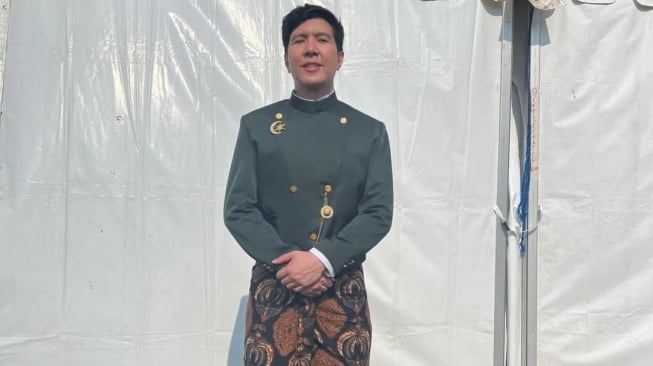 Windah Basudara pembina upacara HUT RI ke-80 mengenakan beskap hijau dan kain batik, berdiri di depan tenda putih.