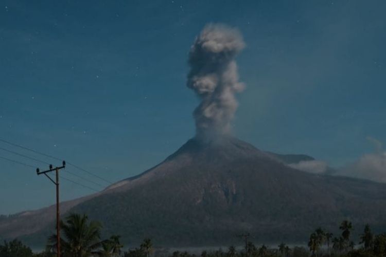 Erupsi Gunung Lewotobi memuntahkan kolom abu vertikal ke langit malam; siluet lereng gunung, pepohonan, dan tiang listrik tampak di latar depan.