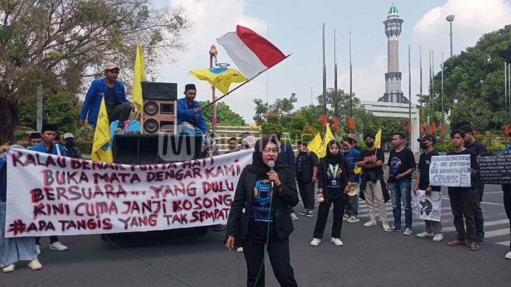 Demo Pati menolak kenaikan PBB di depan pendapa Kabupaten Pati; orator memegang mikrofon, massa membawa spanduk protes dan bendera Merah Putih.