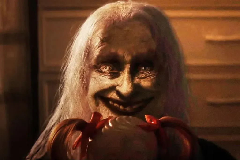 The Conjuring: Last Rites sinopsis — sosok menyeramkan berwajah tua menatap tajam sambil memegang boneka di ruangan gelap